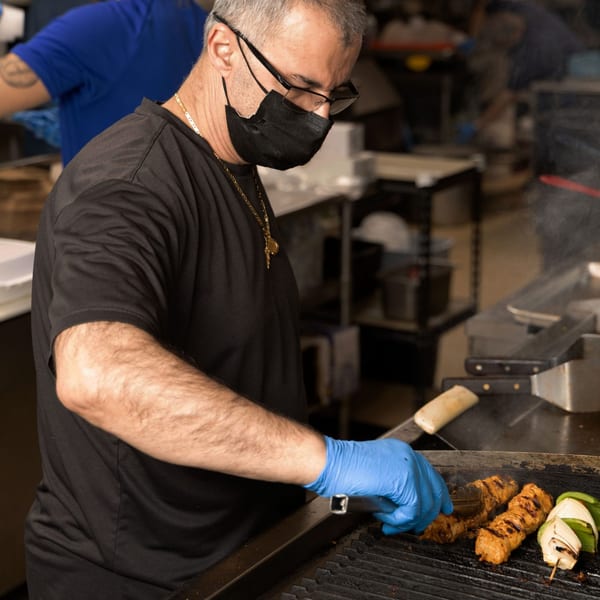 A Yalla Grill chef in a mask and gloves grilling Lebanese shish tawook and vegetables.