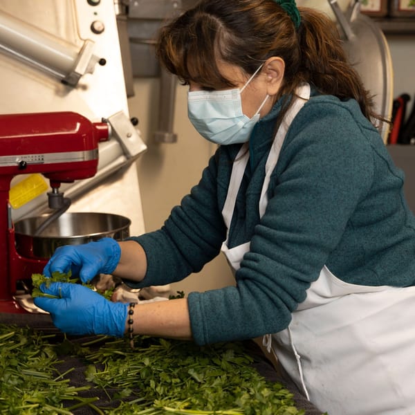 A Yalla Grill chef in a mask and gloves prepares fresh parsley next to a red mixer.