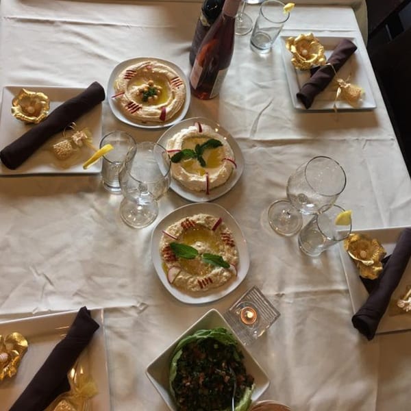 Overhead view of a white tablecloth dining table set with Lebanese hummus, tabbouleh, wine, and place settings.